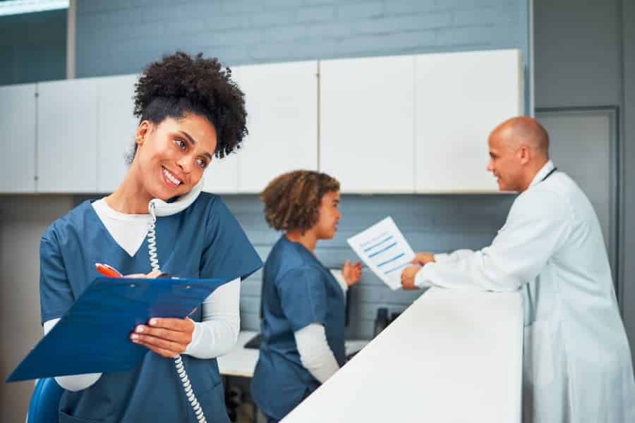 Medical assistant on phone writing notes on clipboard with colleagues in background